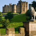Alnwick Castle viewed from Lion Bridge, with a stone lion statue in the foreground and the historic castle rising above green parkland in Northumberland, England.