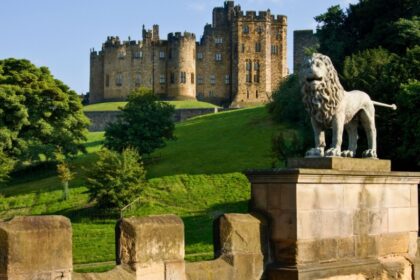 Alnwick Castle viewed from Lion Bridge, with a stone lion statue in the foreground and the historic castle rising above green parkland in Northumberland, England.