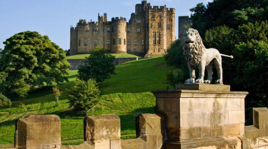 Alnwick Castle viewed from Lion Bridge, with a stone lion statue in the foreground and the historic castle rising above green parkland in Northumberland, England.