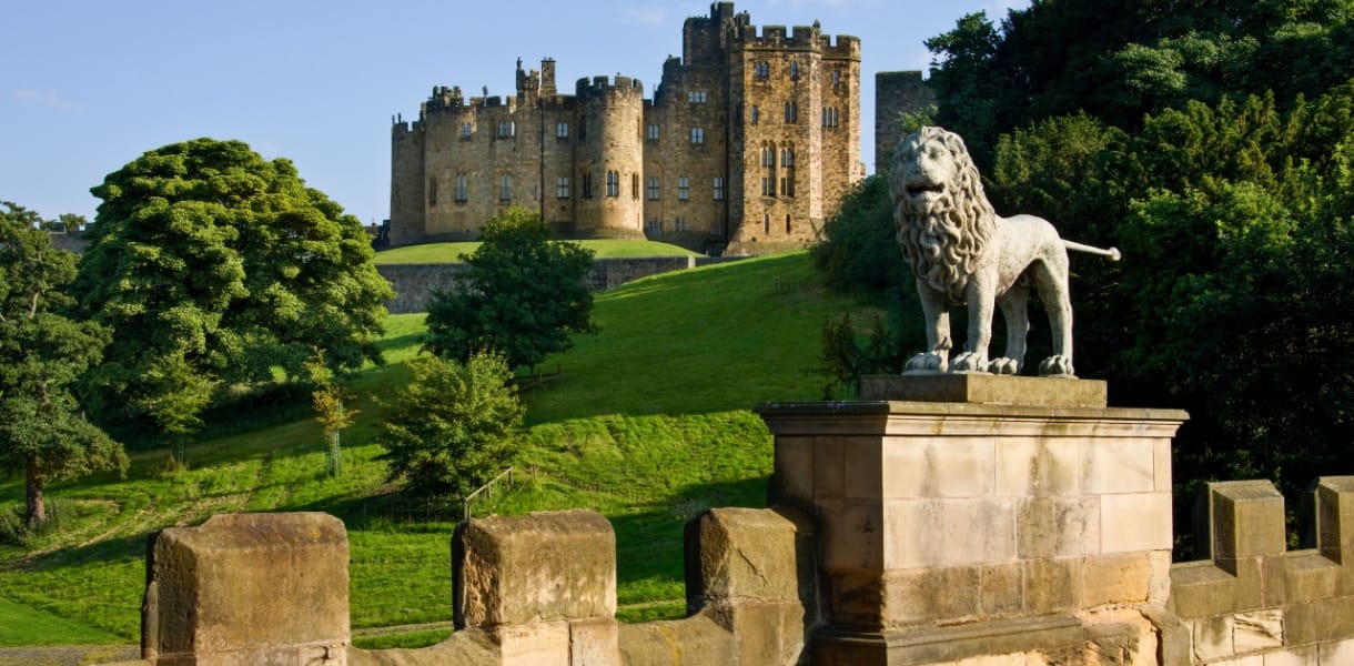 Alnwick Castle viewed from Lion Bridge, with a stone lion statue in the foreground and the historic castle rising above green parkland in Northumberland, England.