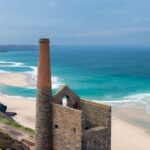 The ruins of a Cornish tin mine standing on a cliff, a wide sweeping beach in the background with a turquoise sea and clear blue sky