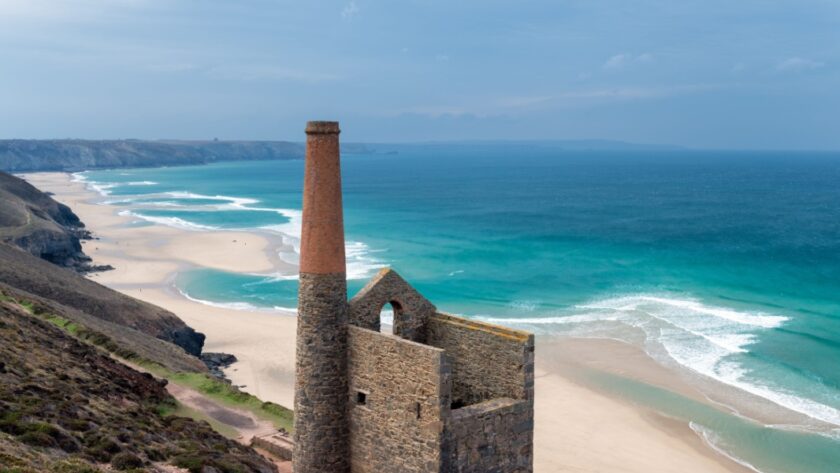 The ruins of a Cornish tin mine standing on a cliff, a wide sweeping beach in the background with a turquoise sea and clear blue sky
