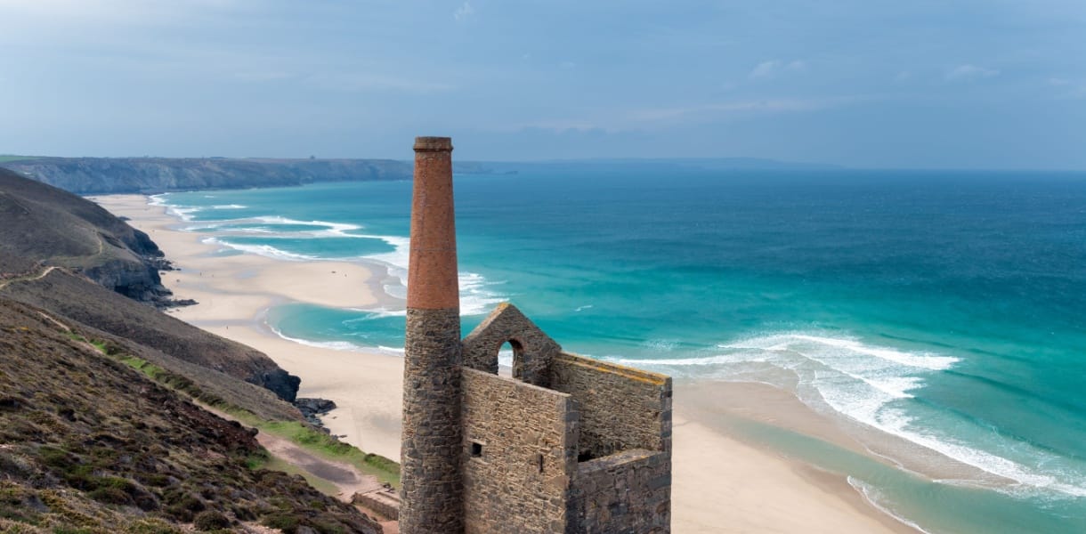 The ruins of a Cornish tin mine standing on a cliff, a wide sweeping beach in the background with a turquoise sea and clear blue sky