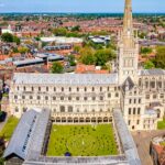Aerial view of Norwich Cathedral with its tall spire, surrounded by the city’s red-roofed buildings, green lawns and trees under a clear blue sky.