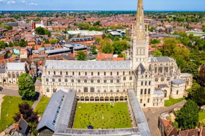 Aerial view of Norwich Cathedral with its tall spire, surrounded by the city’s red-roofed buildings, green lawns and trees under a clear blue sky.