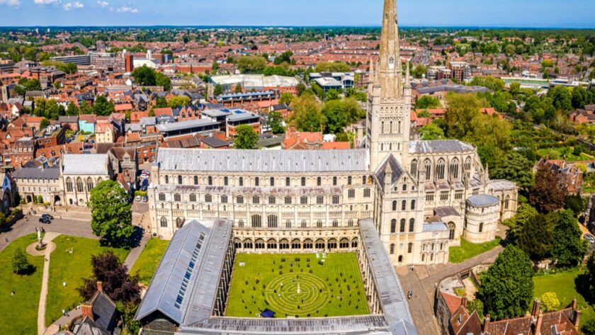 Aerial view of Norwich Cathedral with its tall spire, surrounded by the city’s red-roofed buildings, green lawns and trees under a clear blue sky.