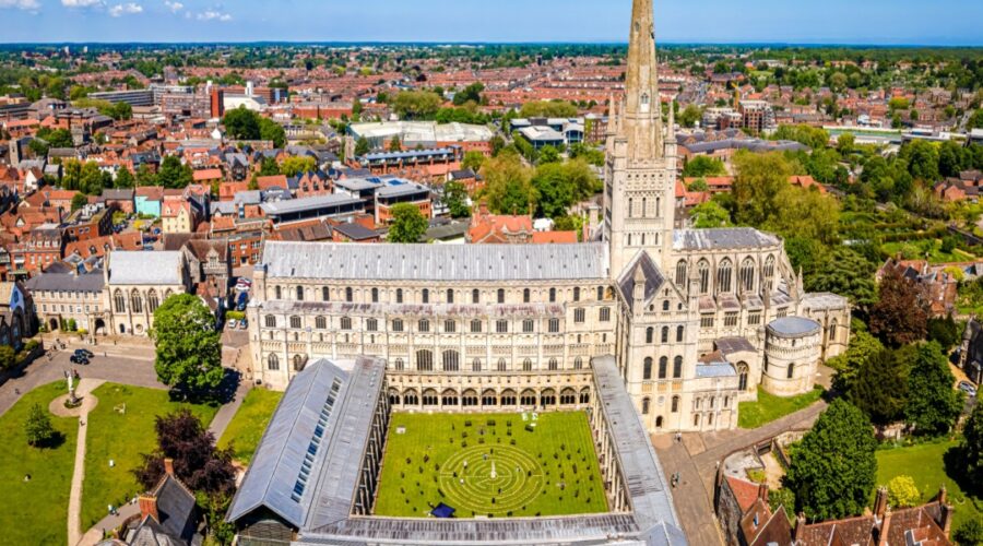 Aerial view of Norwich Cathedral with its tall spire, surrounded by the city’s red-roofed buildings, green lawns and trees under a clear blue sky.