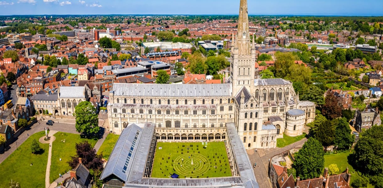 Aerial view of Norwich Cathedral with its tall spire, surrounded by the city’s red-roofed buildings, green lawns and trees under a clear blue sky.