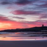 A vivid red sky as the sun rises over the ruins of Dunstanburgh castle with the sky reflected in the wet sand of Embleton Bay beach in the foreground