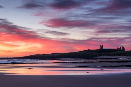 A vivid red sky as the sun rises over the ruins of Dunstanburgh castle with the sky reflected in the wet sand of Embleton Bay beach in the foreground