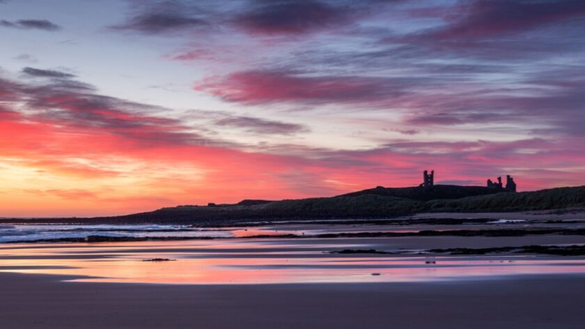 A vivid red sky as the sun rises over the ruins of Dunstanburgh castle with the sky reflected in the wet sand of Embleton Bay beach in the foreground
