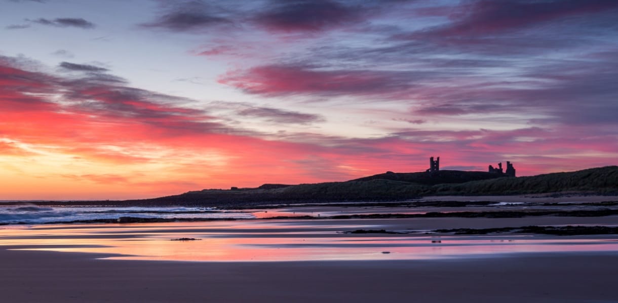 A vivid red sky as the sun rises over the ruins of Dunstanburgh castle with the sky reflected in the wet sand of Embleton Bay beach in the foreground