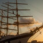 A five masted sailing ship in a harbour with a sunset in the background