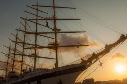 A five masted sailing ship in a harbour with a sunset in the background