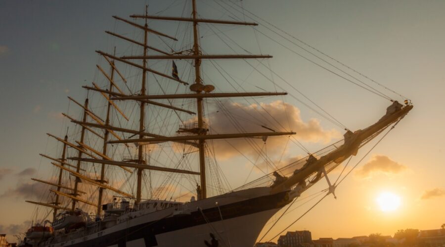 A five masted sailing ship in a harbour with a sunset in the background