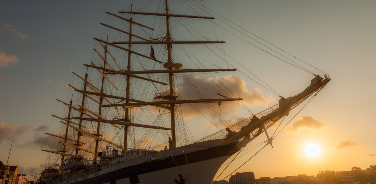 A five masted sailing ship in a harbour with a sunset in the background
