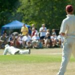 Amateur cricketers in white uniforms play a match on a sunlit village green as spectators watch from under blue gazebos.