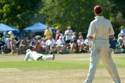 Amateur cricketers in white uniforms play a match on a sunlit village green as spectators watch from under blue gazebos.