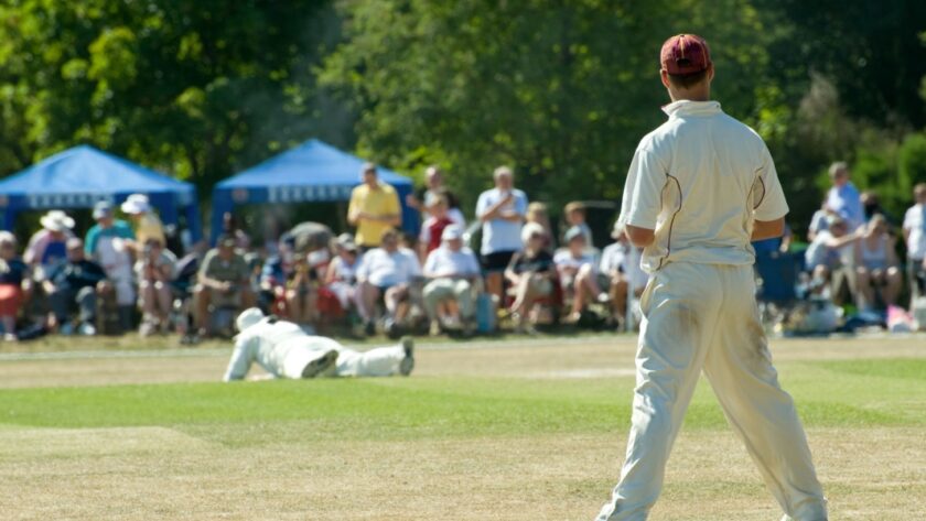 Amateur cricketers in white uniforms play a match on a sunlit village green as spectators watch from under blue gazebos.