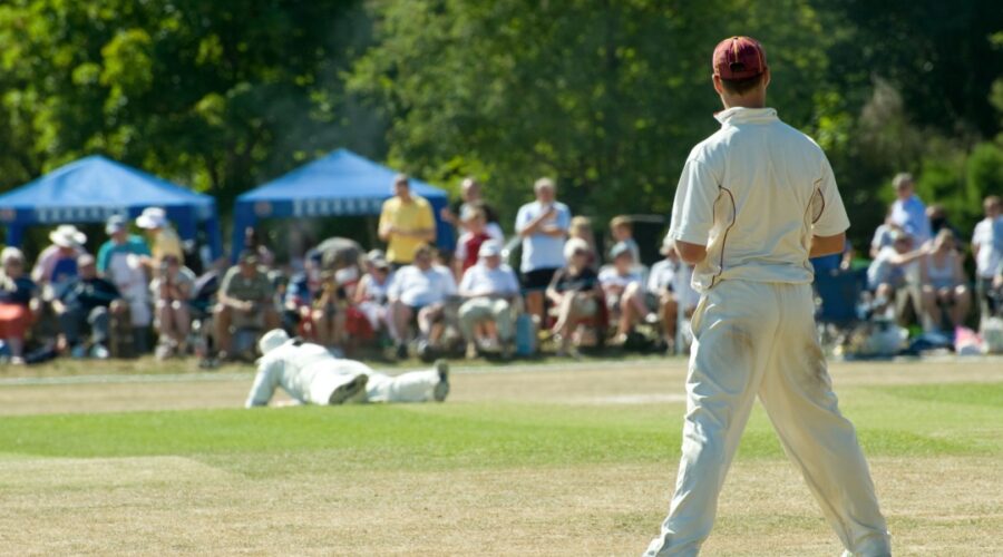 Amateur cricketers in white uniforms play a match on a sunlit village green as spectators watch from under blue gazebos.