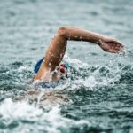 Wild Swimming in South East England.Open-water swimmer in a blue cap and goggles performing a front-crawl stroke, arm outstretched and water spraying amid dark, choppy waves
