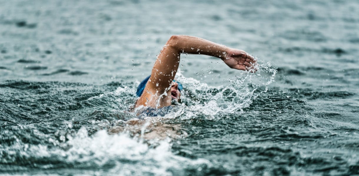 Wild Swimming in South East England.Open-water swimmer in a blue cap and goggles performing a front-crawl stroke, arm outstretched and water spraying amid dark, choppy waves