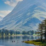 A scenic view of Buttermere in the Lake District, Cumbria, with calm reflective water, people paddling near the shoreline, and a dramatic, sunlit mountain rising steeply in the background. Tall pine trees line the right edge of the lake under a bright blue sky with scattered clouds.