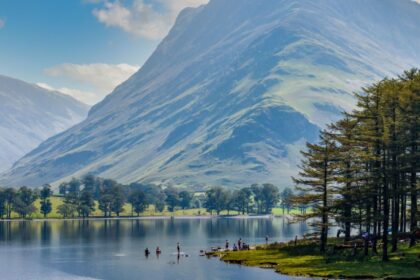 A scenic view of Buttermere in the Lake District, Cumbria, with calm reflective water, people paddling near the shoreline, and a dramatic, sunlit mountain rising steeply in the background. Tall pine trees line the right edge of the lake under a bright blue sky with scattered clouds.