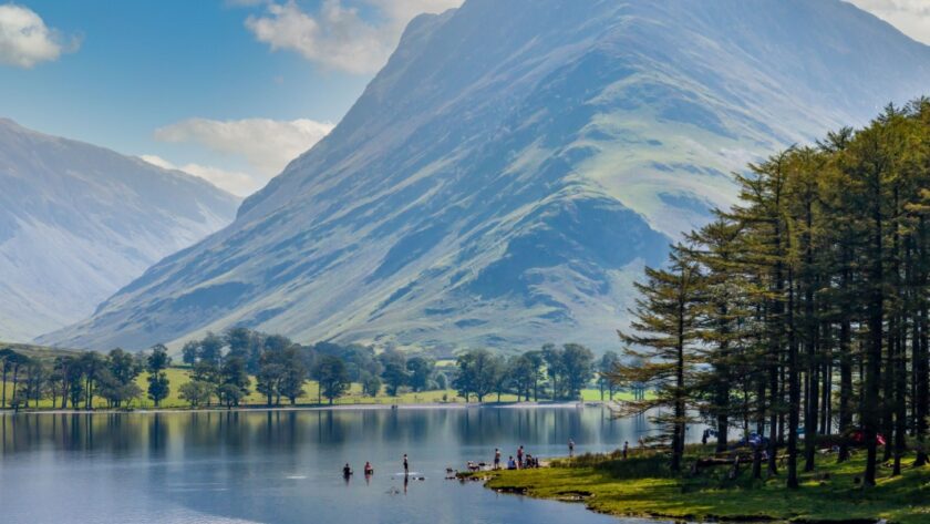 A scenic view of Buttermere in the Lake District, Cumbria, with calm reflective water, people paddling near the shoreline, and a dramatic, sunlit mountain rising steeply in the background. Tall pine trees line the right edge of the lake under a bright blue sky with scattered clouds.