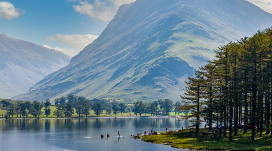 A scenic view of Buttermere in the Lake District, Cumbria, with calm reflective water, people paddling near the shoreline, and a dramatic, sunlit mountain rising steeply in the background. Tall pine trees line the right edge of the lake under a bright blue sky with scattered clouds.