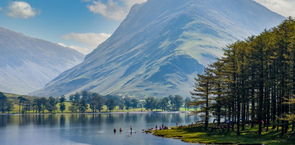 A scenic view of Buttermere in the Lake District, Cumbria, with calm reflective water, people paddling near the shoreline, and a dramatic, sunlit mountain rising steeply in the background. Tall pine trees line the right edge of the lake under a bright blue sky with scattered clouds.