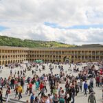 A bustling scene at the Piece Hall in Halifax, West Yorkshire, with hundreds of people gathered in the large open courtyard surrounded by symmetrical stone arcades and columns. In the background, a tall church spire rises above green hills under a partly cloudy sky.