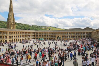 A bustling scene at the Piece Hall in Halifax, West Yorkshire, with hundreds of people gathered in the large open courtyard surrounded by symmetrical stone arcades and columns. In the background, a tall church spire rises above green hills under a partly cloudy sky.