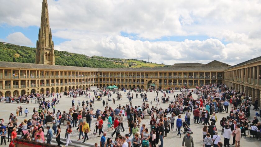 A bustling scene at the Piece Hall in Halifax, West Yorkshire, with hundreds of people gathered in the large open courtyard surrounded by symmetrical stone arcades and columns. In the background, a tall church spire rises above green hills under a partly cloudy sky.
