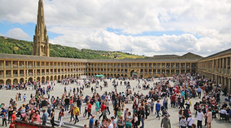 A bustling scene at the Piece Hall in Halifax, West Yorkshire, with hundreds of people gathered in the large open courtyard surrounded by symmetrical stone arcades and columns. In the background, a tall church spire rises above green hills under a partly cloudy sky.