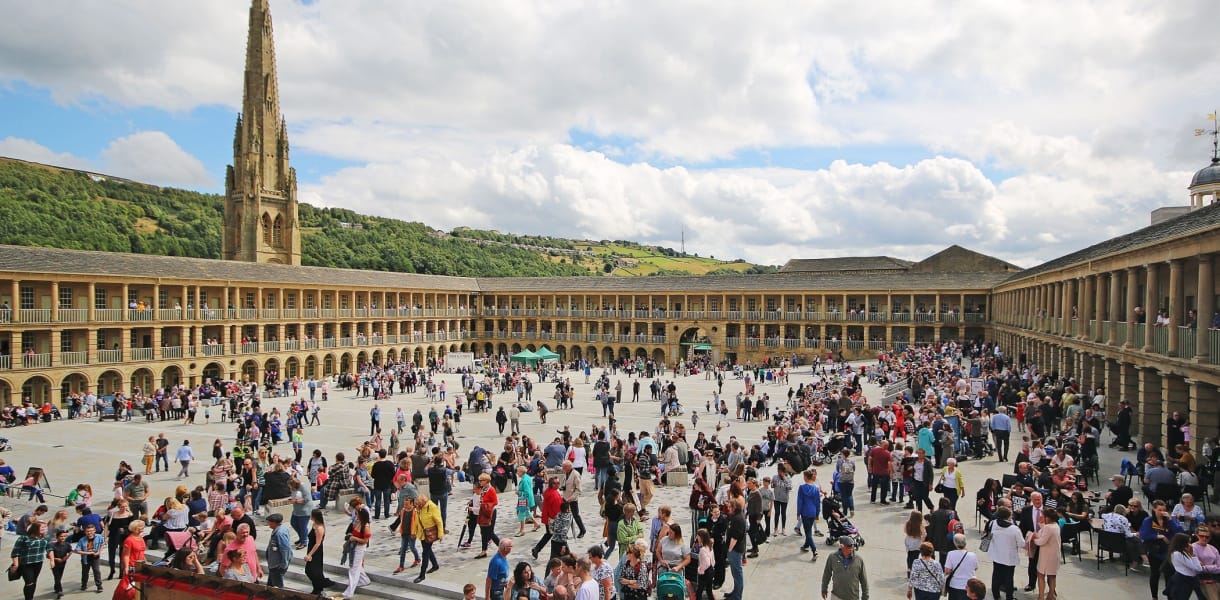 A bustling scene at the Piece Hall in Halifax, West Yorkshire, with hundreds of people gathered in the large open courtyard surrounded by symmetrical stone arcades and columns. In the background, a tall church spire rises above green hills under a partly cloudy sky.
