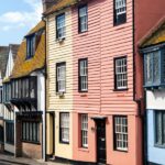 Colourful historic buildings line a narrow street in Hastings Old Town, East Sussex, with a glimpse of the sea and a bright blue building visible at the end of the road under a sunny sky