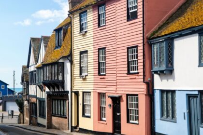 Colourful historic buildings line a narrow street in Hastings Old Town, East Sussex, with a glimpse of the sea and a bright blue building visible at the end of the road under a sunny sky