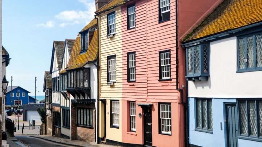Colourful historic buildings line a narrow street in Hastings Old Town, East Sussex, with a glimpse of the sea and a bright blue building visible at the end of the road under a sunny sky