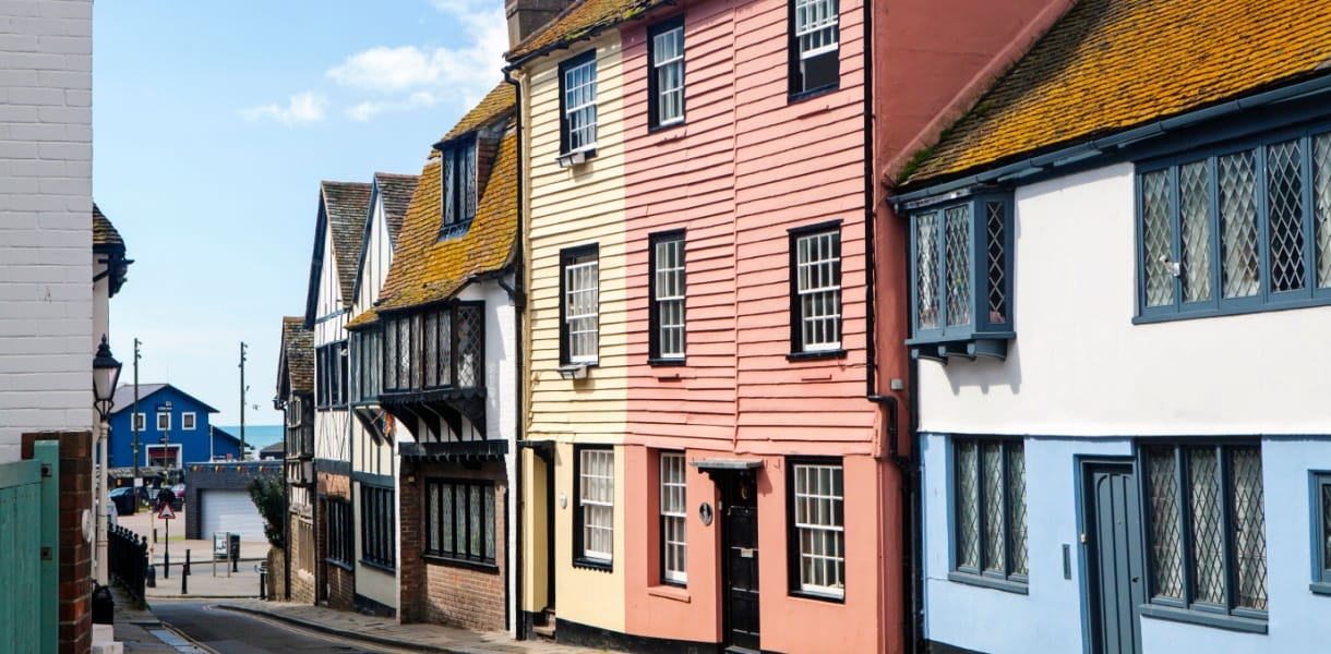 Colourful historic buildings line a narrow street in Hastings Old Town, East Sussex, with a glimpse of the sea and a bright blue building visible at the end of the road under a sunny sky