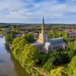 Aerial view of Holy Trinity Church in Stratford-upon-Avon, Warwickshire, surrounded by lush green trees and situated beside the calm River Avon, with the town and rolling countryside stretching into the distance under a blue sky.