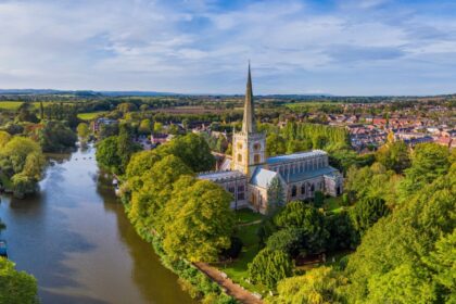 Aerial view of Holy Trinity Church in Stratford-upon-Avon, Warwickshire, surrounded by lush green trees and situated beside the calm River Avon, with the town and rolling countryside stretching into the distance under a blue sky.