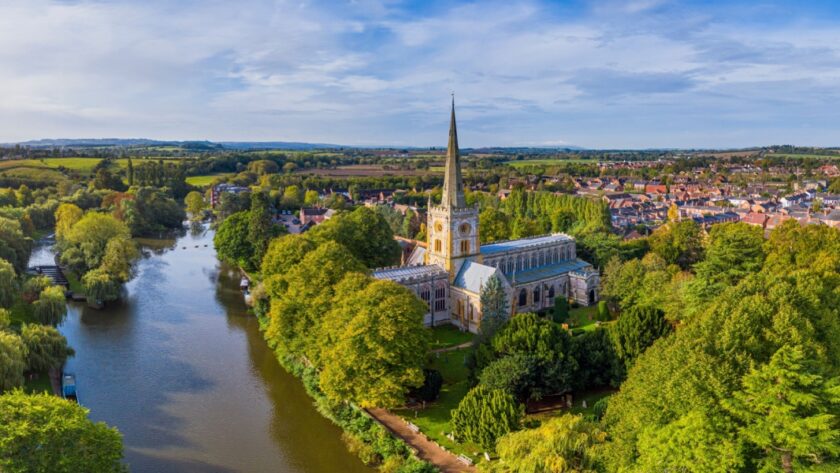 Aerial view of Holy Trinity Church in Stratford-upon-Avon, Warwickshire, surrounded by lush green trees and situated beside the calm River Avon, with the town and rolling countryside stretching into the distance under a blue sky.