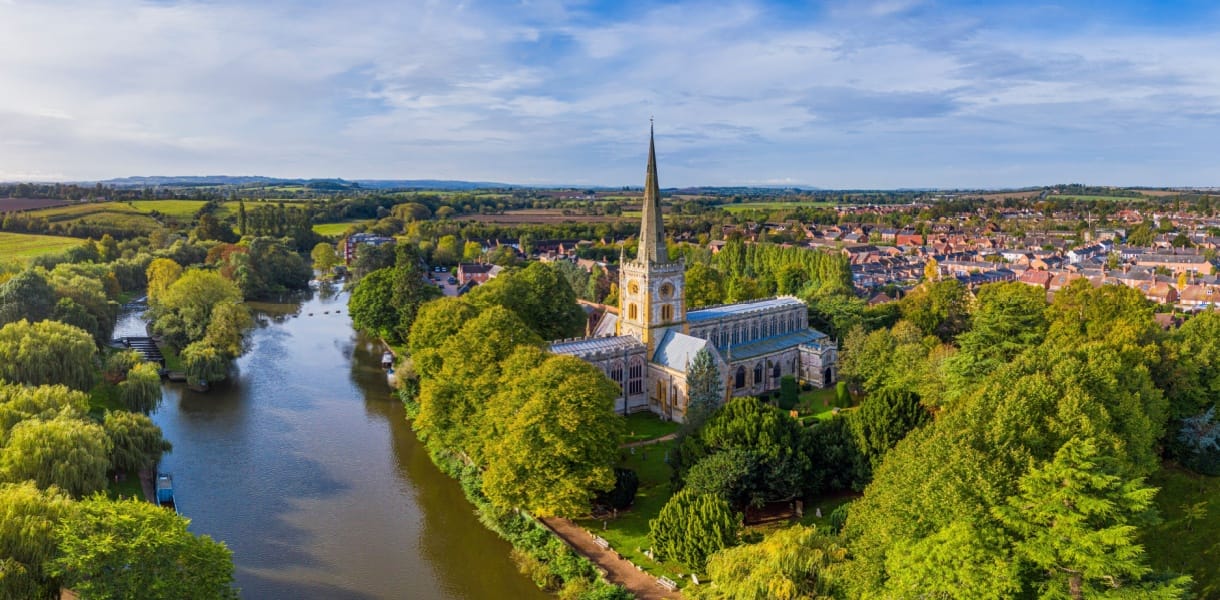 Aerial view of Holy Trinity Church in Stratford-upon-Avon, Warwickshire, surrounded by lush green trees and situated beside the calm River Avon, with the town and rolling countryside stretching into the distance under a blue sky.