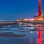 Blackpool Tower illuminated at night, reflected in the wet sand of the beach, with the pier lit up in the distance on the Lancashire coast.