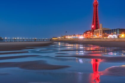 Blackpool Tower illuminated at night, reflected in the wet sand of the beach, with the pier lit up in the distance on the Lancashire coast.