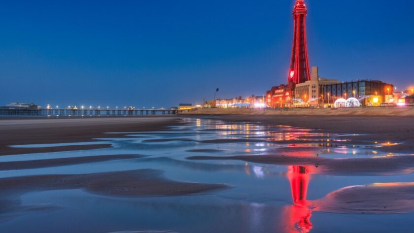 Blackpool Tower illuminated at night, reflected in the wet sand of the beach, with the pier lit up in the distance on the Lancashire coast.