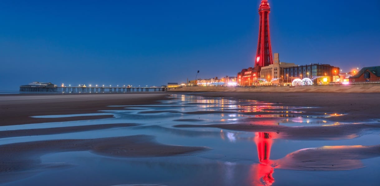 Blackpool Tower illuminated at night, reflected in the wet sand of the beach, with the pier lit up in the distance on the Lancashire coast.