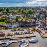 A scenic aerial view of Lymington harbour, showing moored boats and a waterfront lined with traditional buildings. Behind, rows of houses and green trees stretch into the distance under a partly cloudy sky