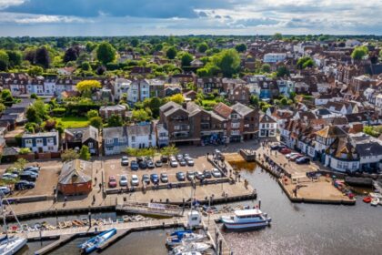 A scenic aerial view of Lymington harbour, showing moored boats and a waterfront lined with traditional buildings. Behind, rows of houses and green trees stretch into the distance under a partly cloudy sky